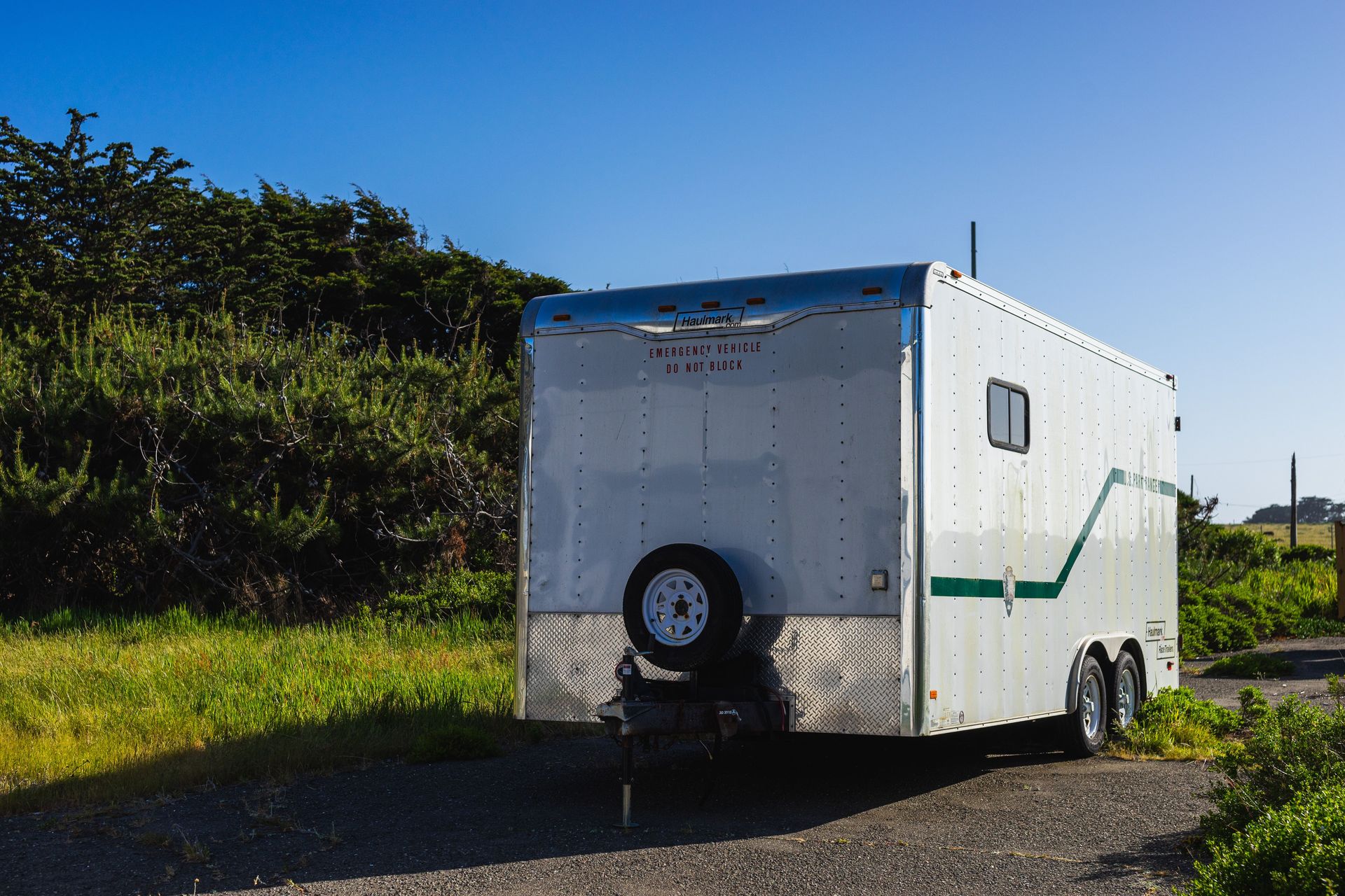 A parked white trailer surrounded by greenery under a clear blue sky in a rural area.