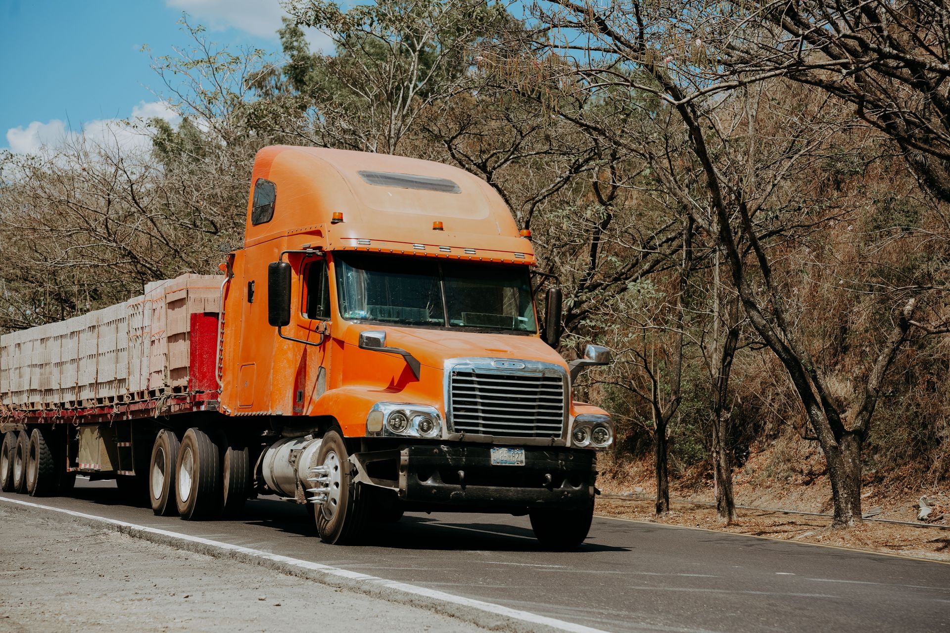 A vibrant orange Freightliner truck drives on a highway through a dry, scenic landscape in El Salvador.